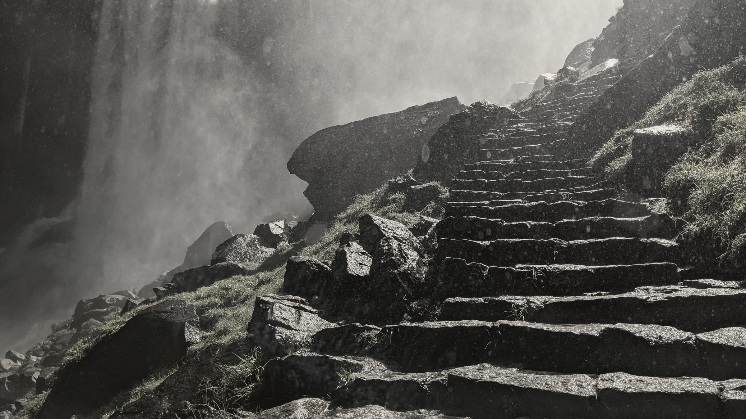 Rough stone steps climbing steeply beside a waterfall, its spray drifting across the rocks in the light.