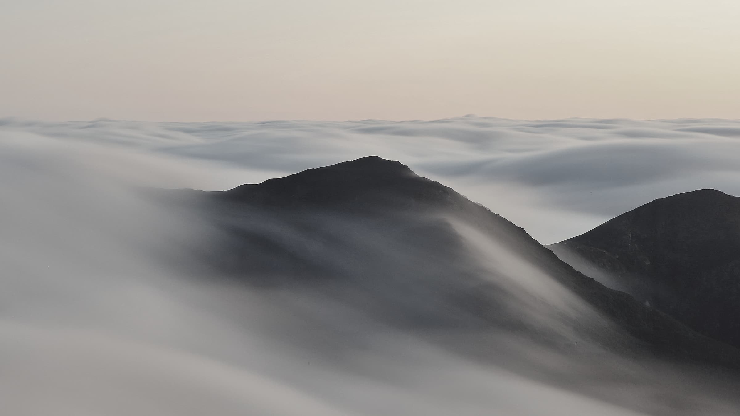 A dark summit standing above a sea of cloud that pours slowly over its shoulders, under a pale dawn sky.