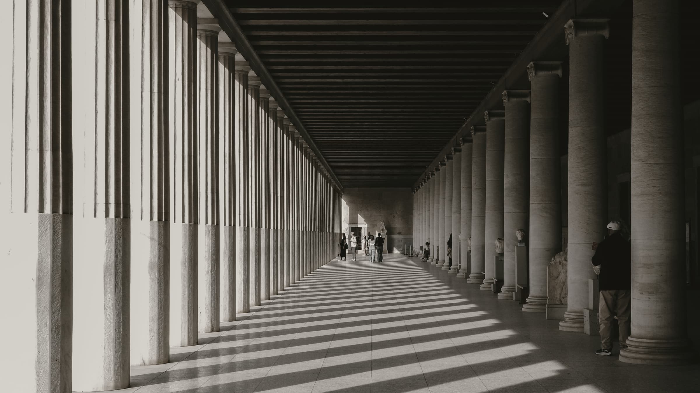 A long stone colonnade in Athens, its fluted columns casting bars of light and shadow across a polished floor.